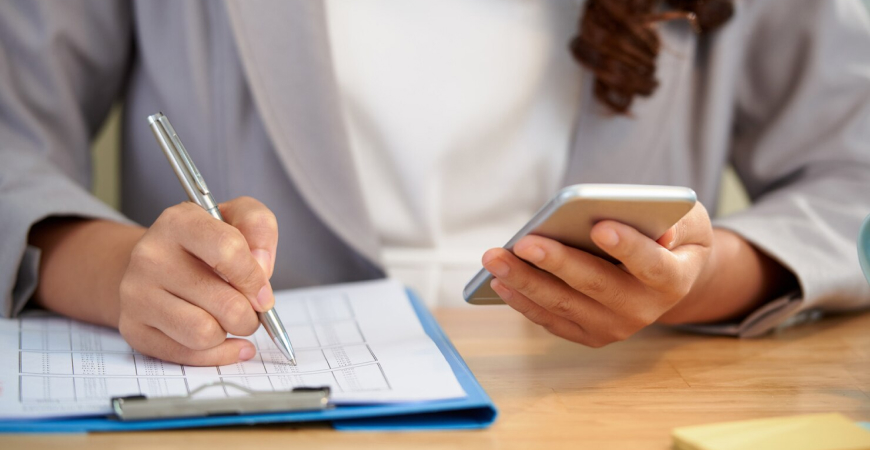Reta final do ano organize obrigações — empresária anotando em prancheta e consultando celular para planejar fechamento do exercício